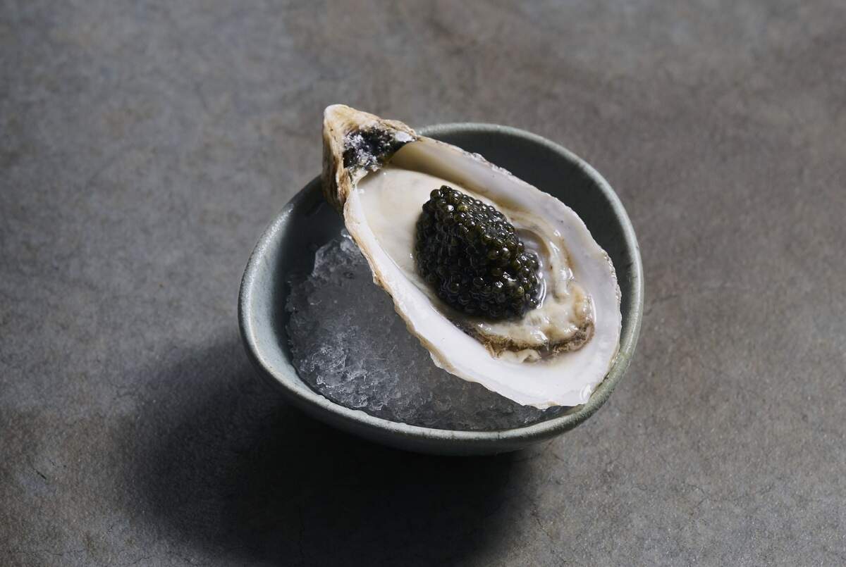 high-angle, close-up shot of a single fresh oyster served on the half shell, presented as a gourmet delicacy. The oyster rests at a slight angle on a bed of finely crushed ice inside a small, rustic, organic-shaped ceramic bowl with a matte, sage green finish. Nestled perfectly inside the glistening oyster shell, a generous spoonful of glossy, dark green-to-black caviar serves as the centerpiece.